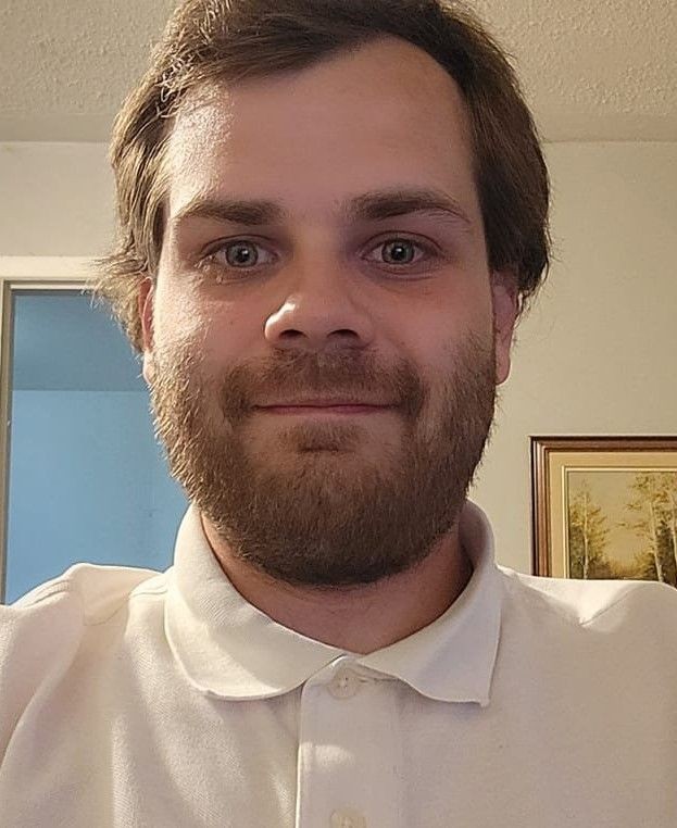 Person wearing a white polo shirt standing indoors with a framed picture and a door in the background.